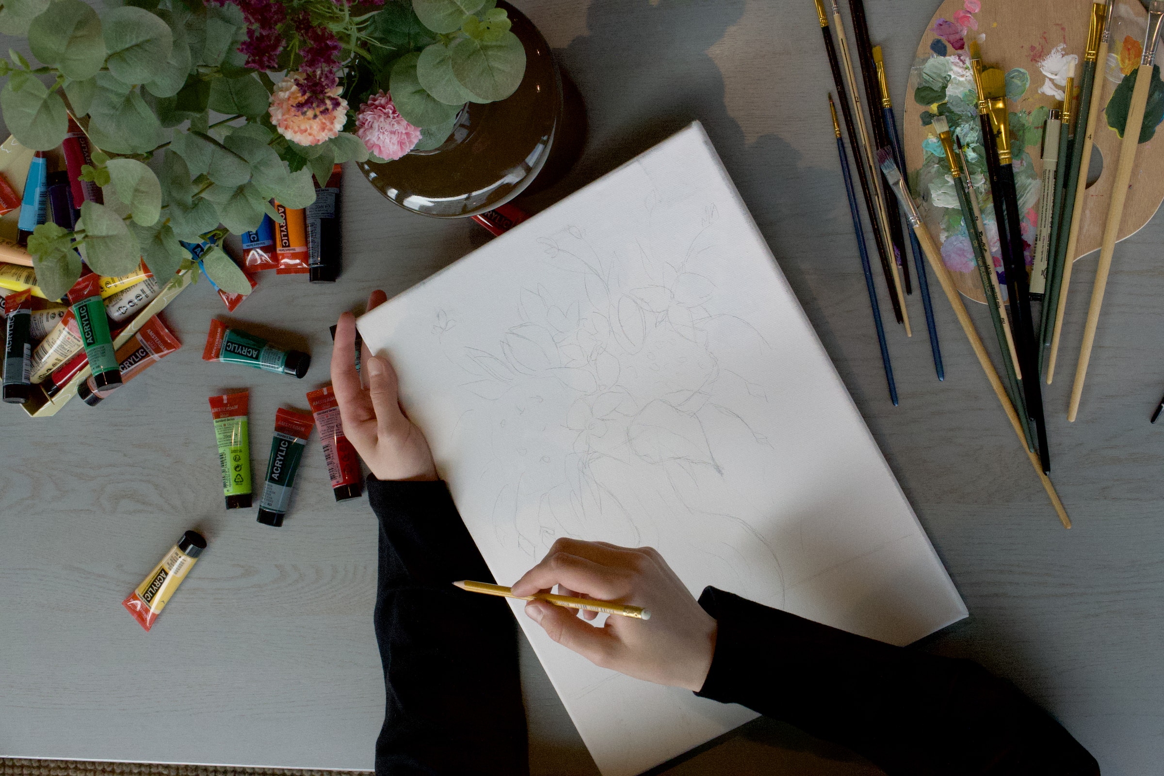 Overhead view — hands holding a canvas with a pencil sketch, paints and brushes arranged around it